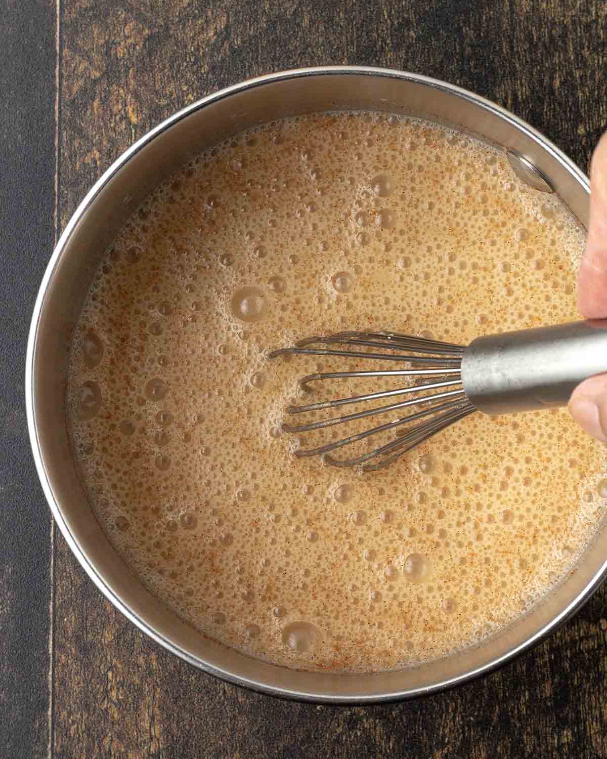 A hand using a metal whisk to mix an oat milk pumpkin spice latte in a small pot.