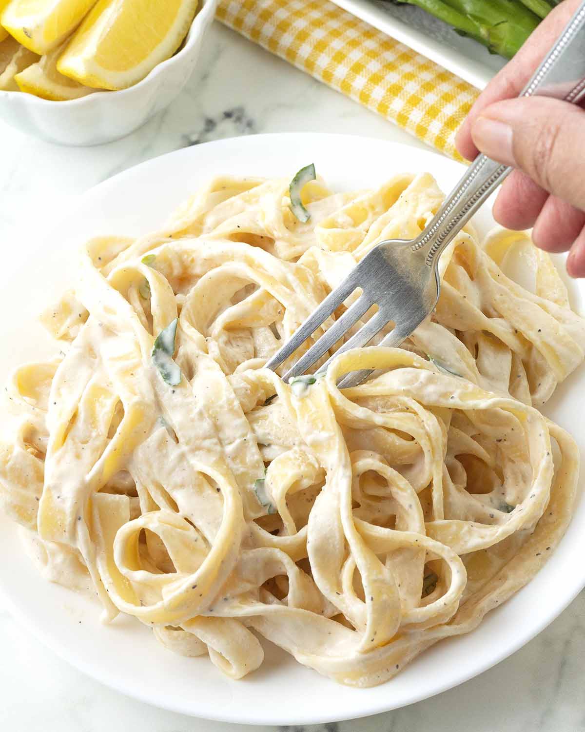 A hand holding a fork, the fork is digging into a plate of pasta with tahini sauce.