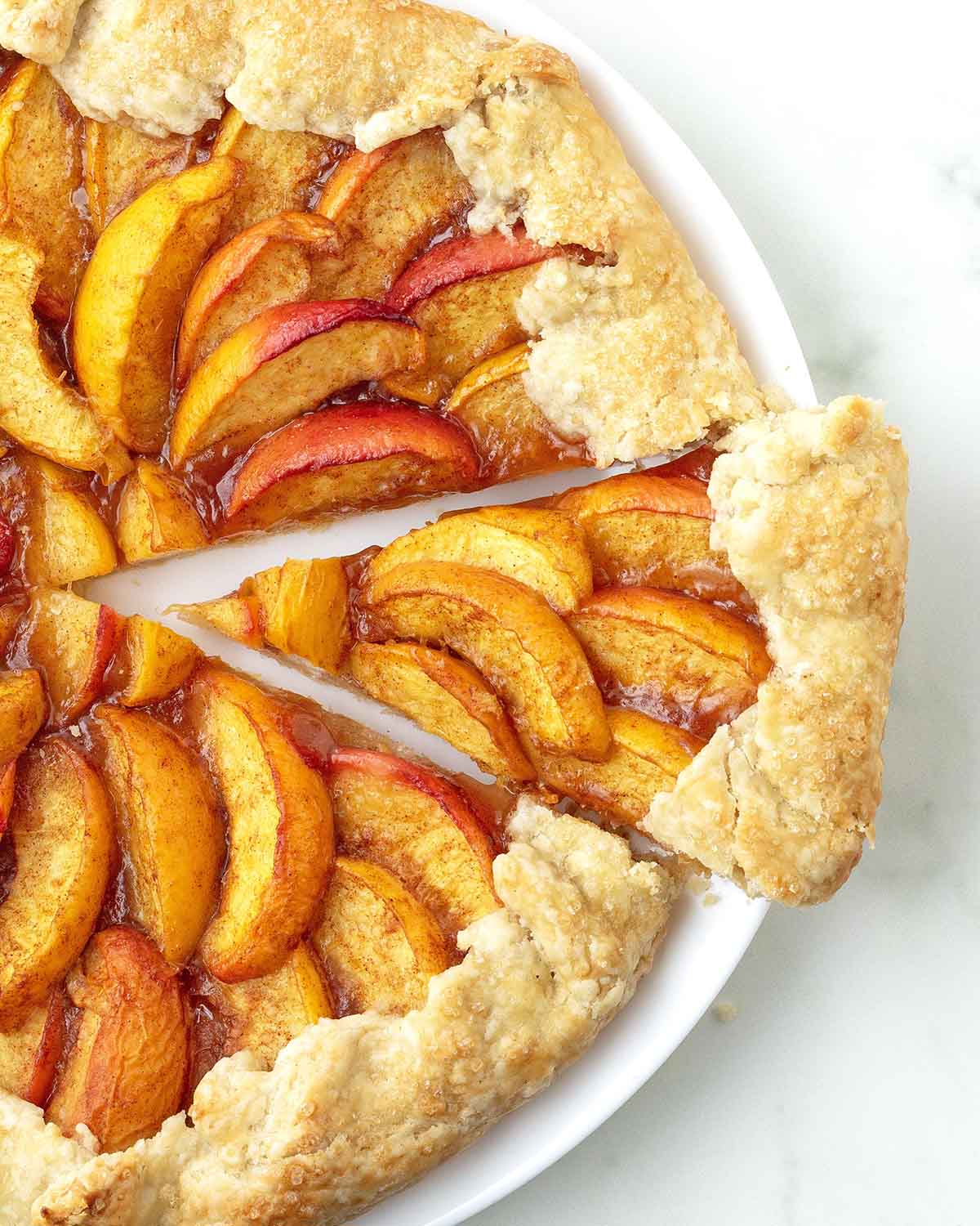 An overhead shot of a fresh peach galette, a slice has been cut and it’s slightly pulled away from the rest of the galette.
