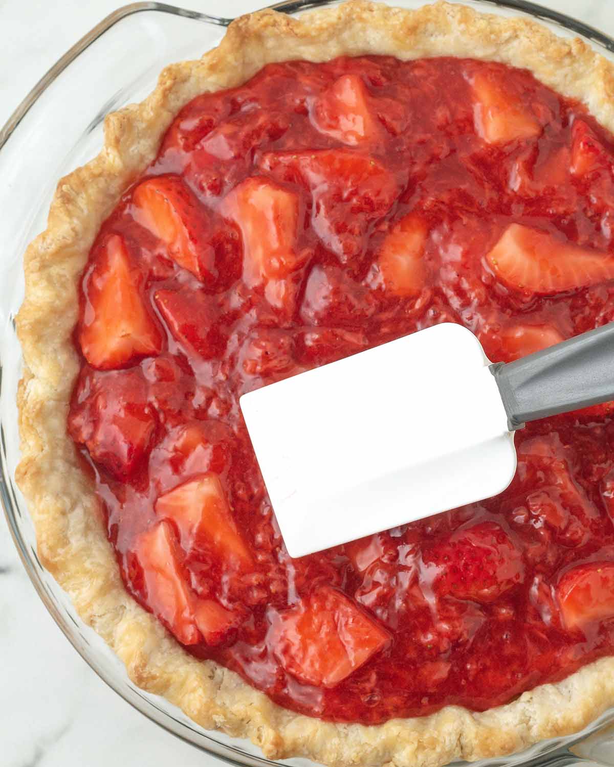 An overhead shot of a strawberry pie in a glass pie dish, a rubber spatula is smoothing out the top of the pie.