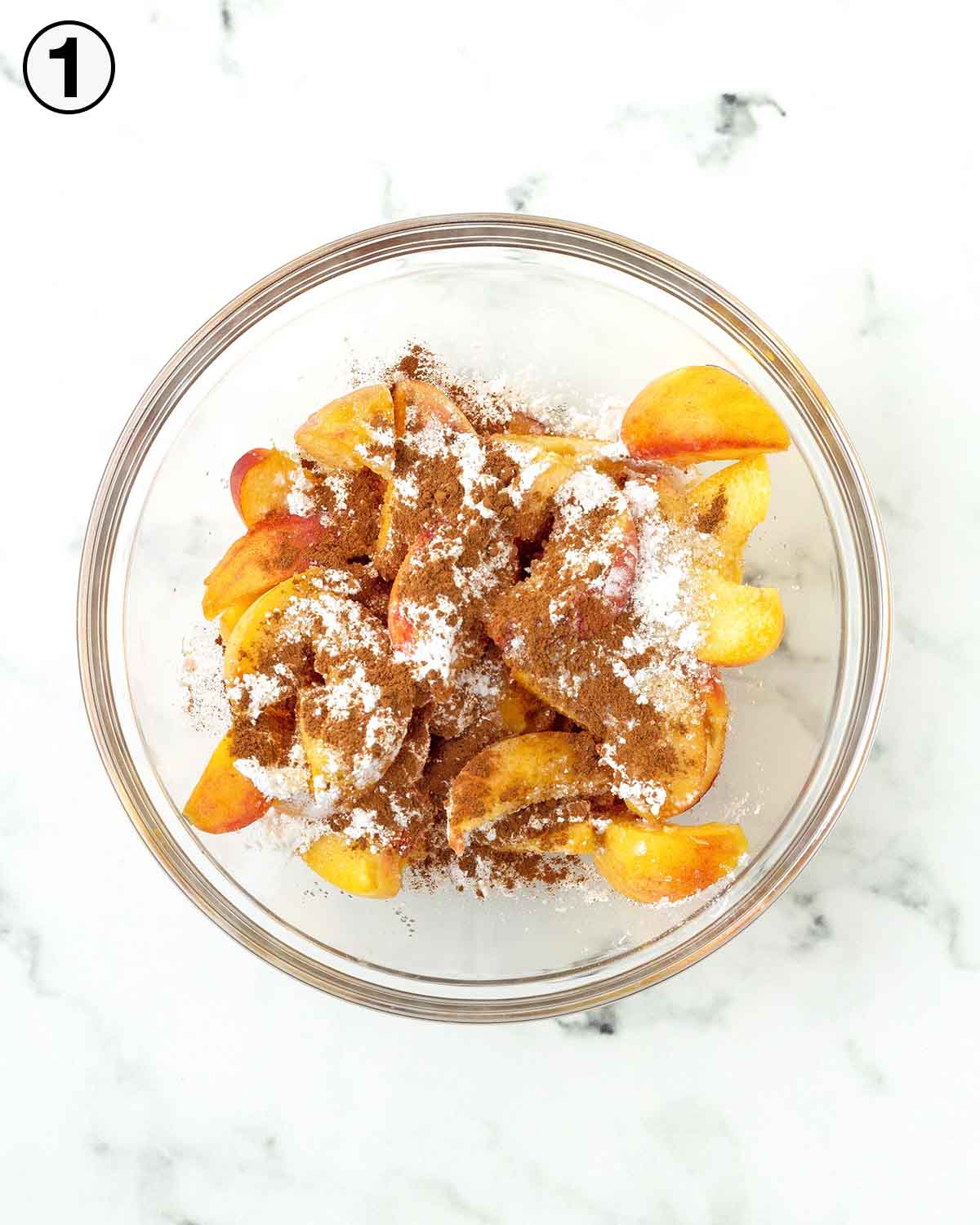 A glass bowl filled with sliced fresh peaches, sugar, cornstarch and cinnamon sits on top of the peaches.
