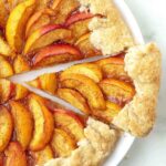 An overhead shot of a fresh peach galette, a slice has been cut and it’s slightly pulled away from the rest of the galette.
