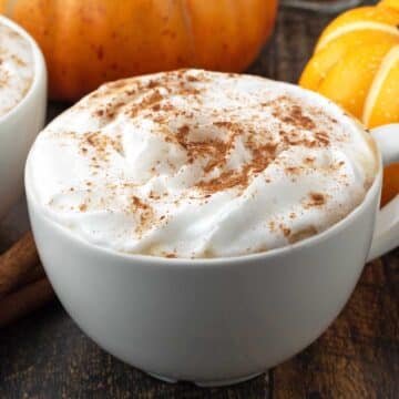 A vegan pumpkin spice latte in a white mug sitting on a dark wood table.