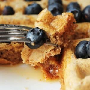 A fork taking away a piece of waffle that is topped with syrup fresh blueberries.