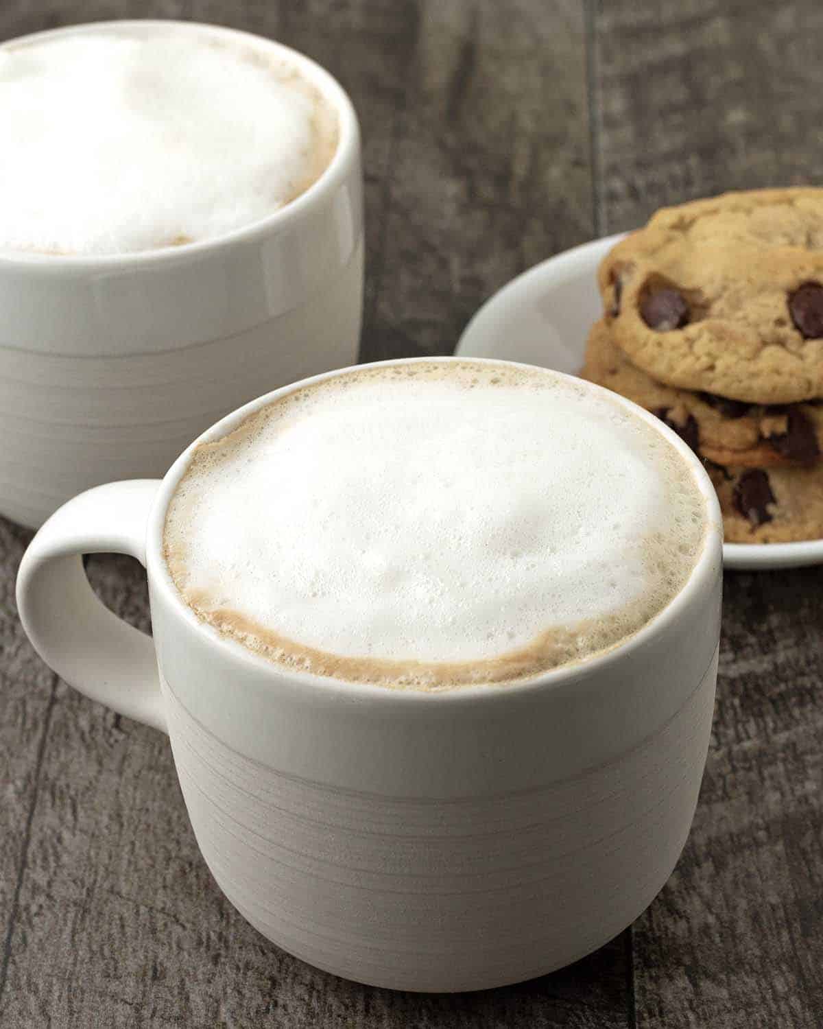 Two hot almond milk lattes in mugs on a table, a dish of cookies sit behind the mugs.