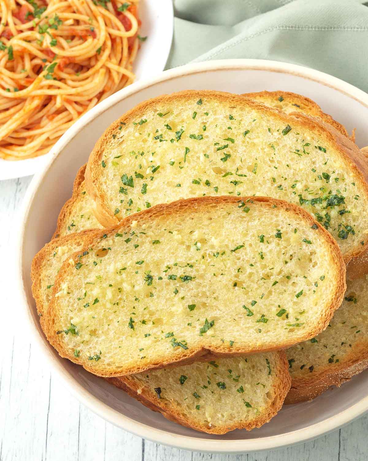 A dish of freshly made vegan garlic bread slices, a plate of spaghetti sits behind the bowl.