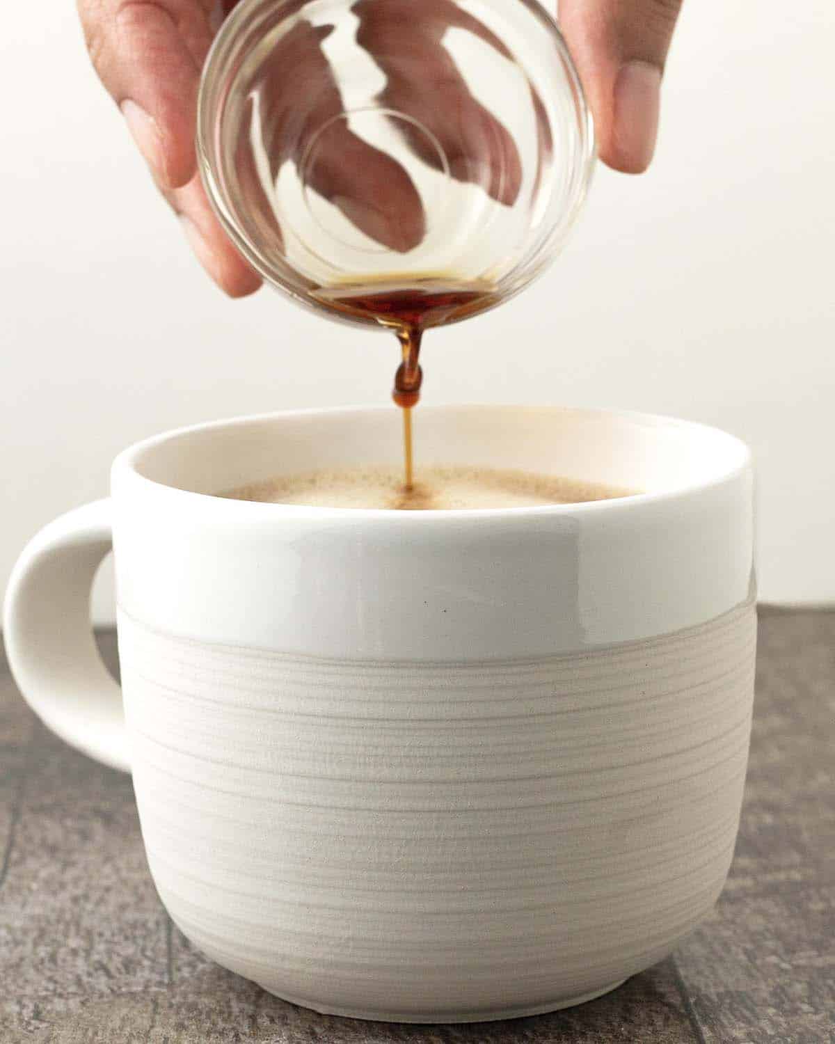 A hand pouring vanilla extract from a small glass bowl into a mug.