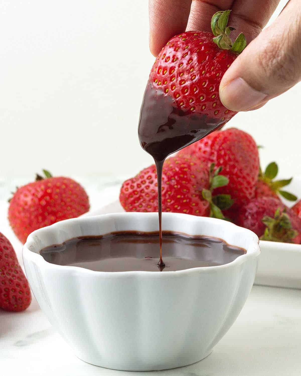 A hand dipping a fresh strawberry into a small bowl of chocolate sauce.