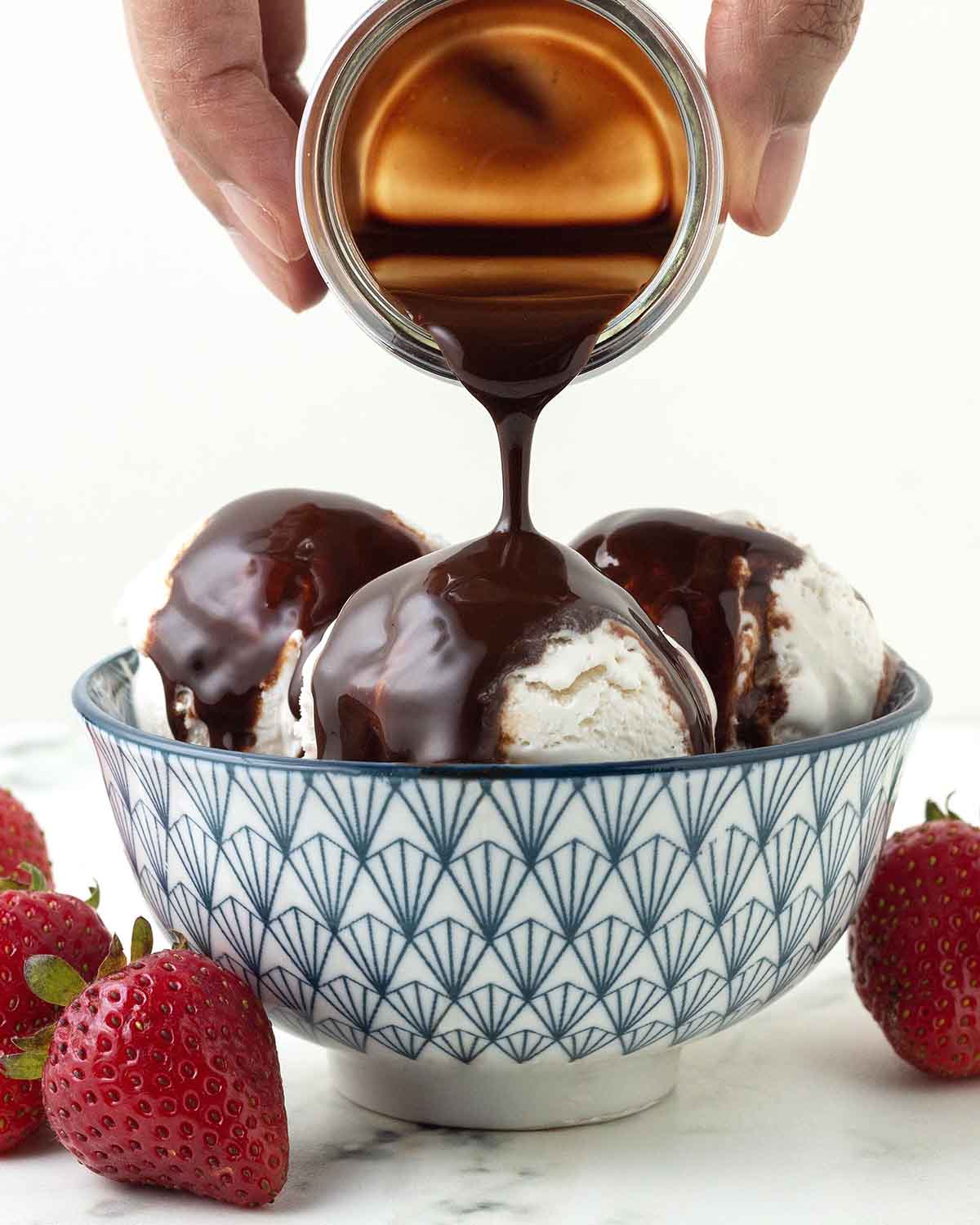 A hand pouring chocolate syrup from a small glass jar onto ice cream in a blue bowl.