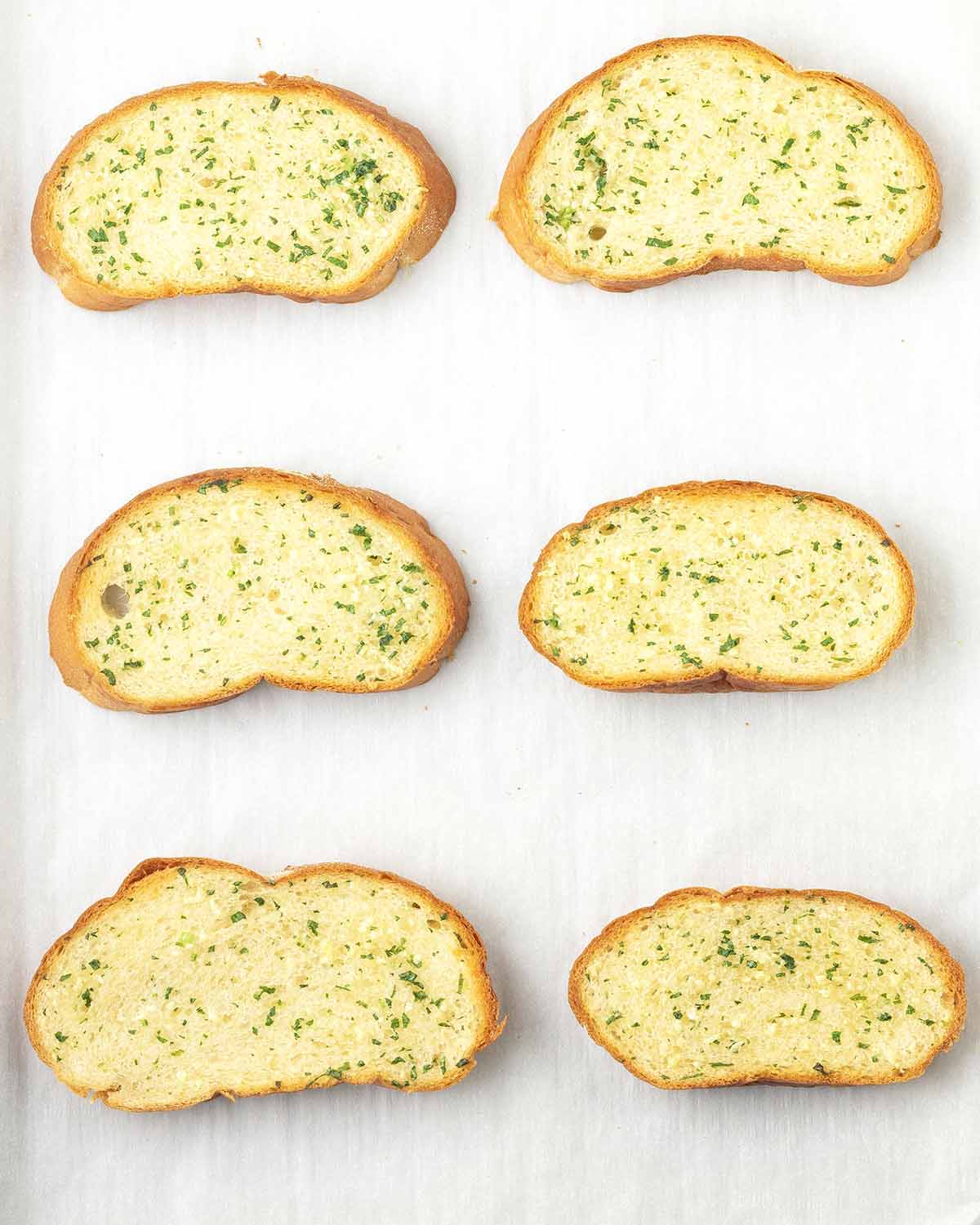 An overhead image showing six slices of freshly baked garlic bread on a parchment lined baking pan.