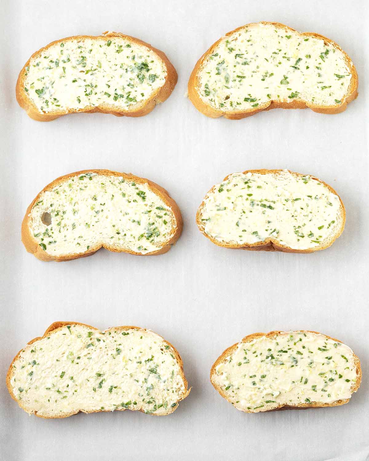 An overhead image showing six slices of bread with garlic spread on a parchment lined baking pan before it is toasted.