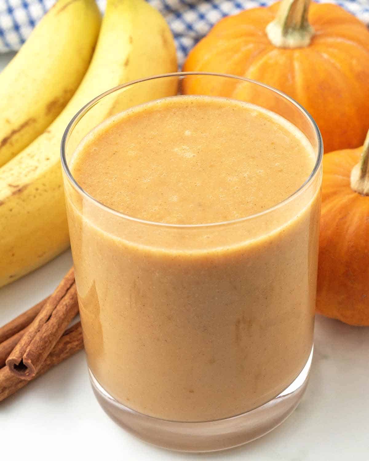A glass of filled with almond milk pumpkin smoothie, bananas, mini pumpkins, and cinnamon sticks sit around the glass.