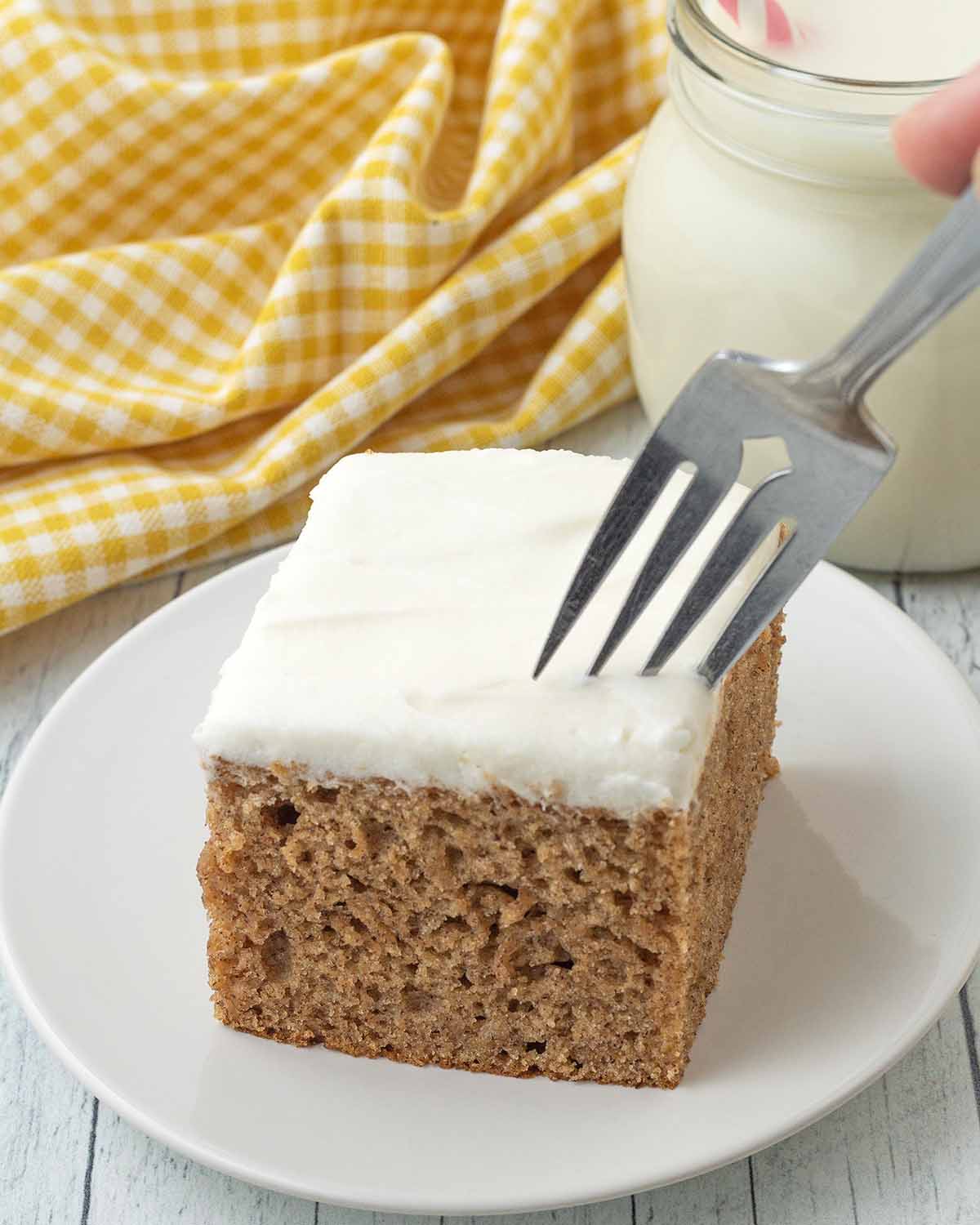 A hand holding a fork that is digging into a square of frosted vegan banana cake.