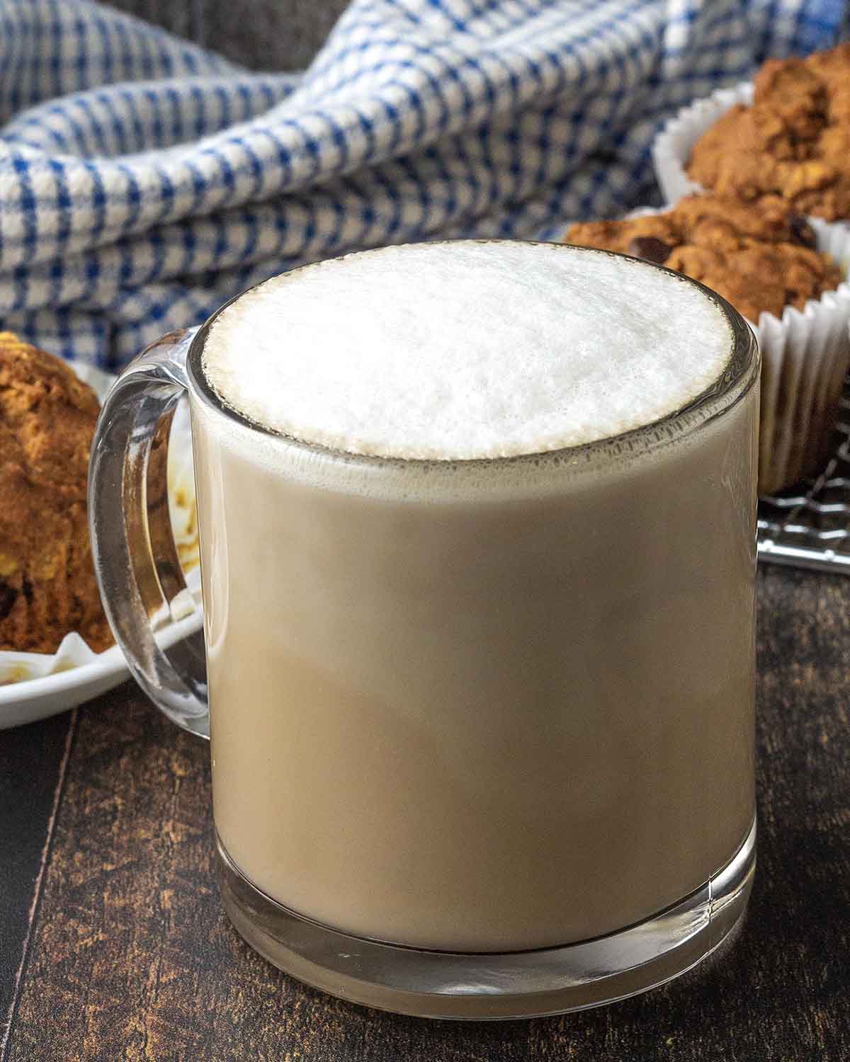 An oat latte in a glass mug sitting on a dark wood table, muffins sit behind the mug.