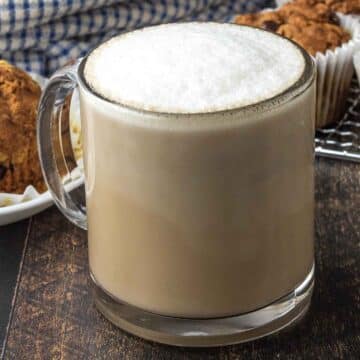 An oat milk latte in a glass mug sitting on a dark wood table.