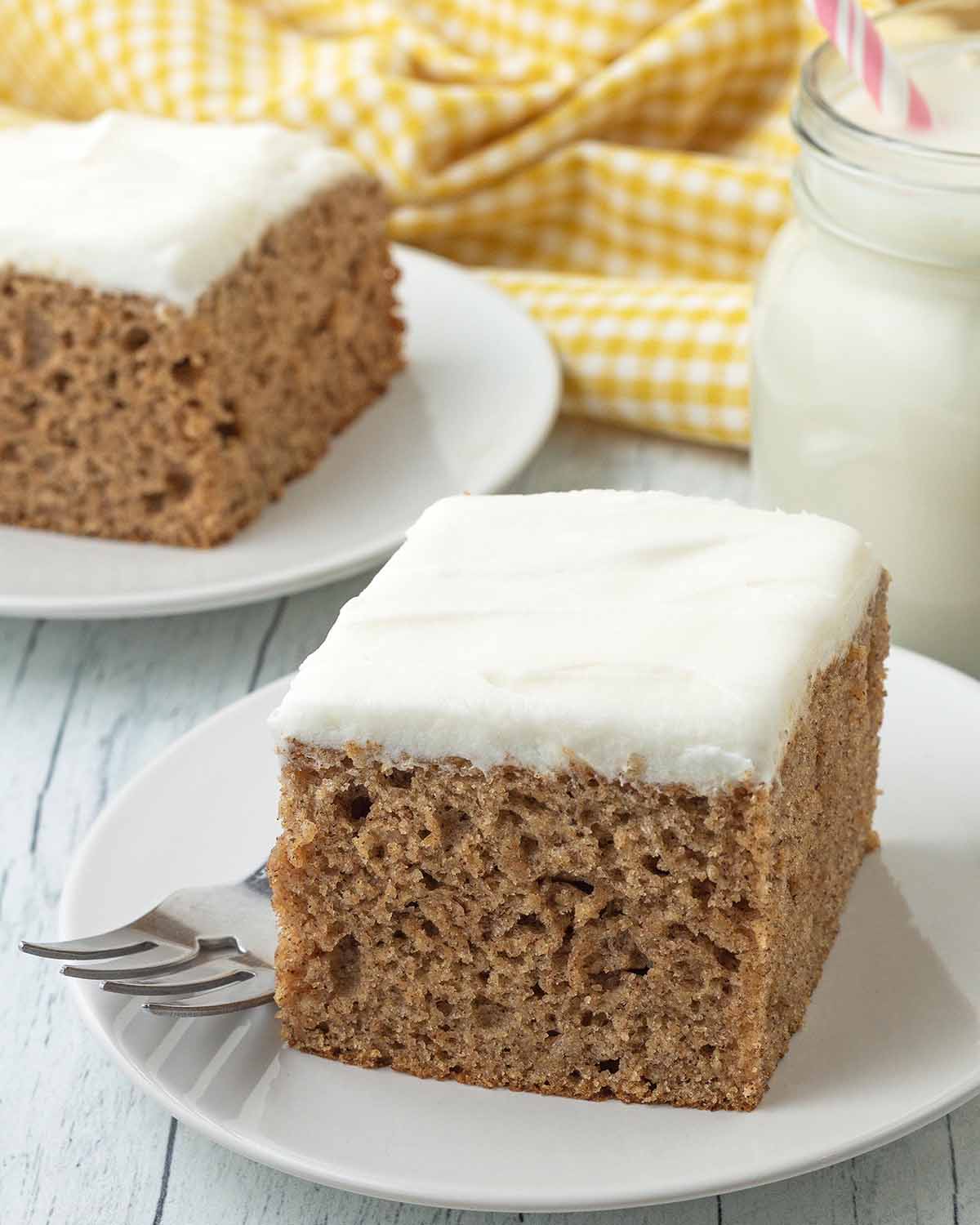 A square piece of banana cake with frosting sitting on a small white plate with a fork on the side.