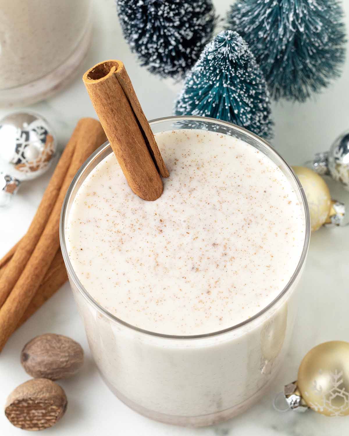 An overhead image of almond milk eggnog in a glass, the glass has a cinnamon stick in it and is surrounded by decorations.