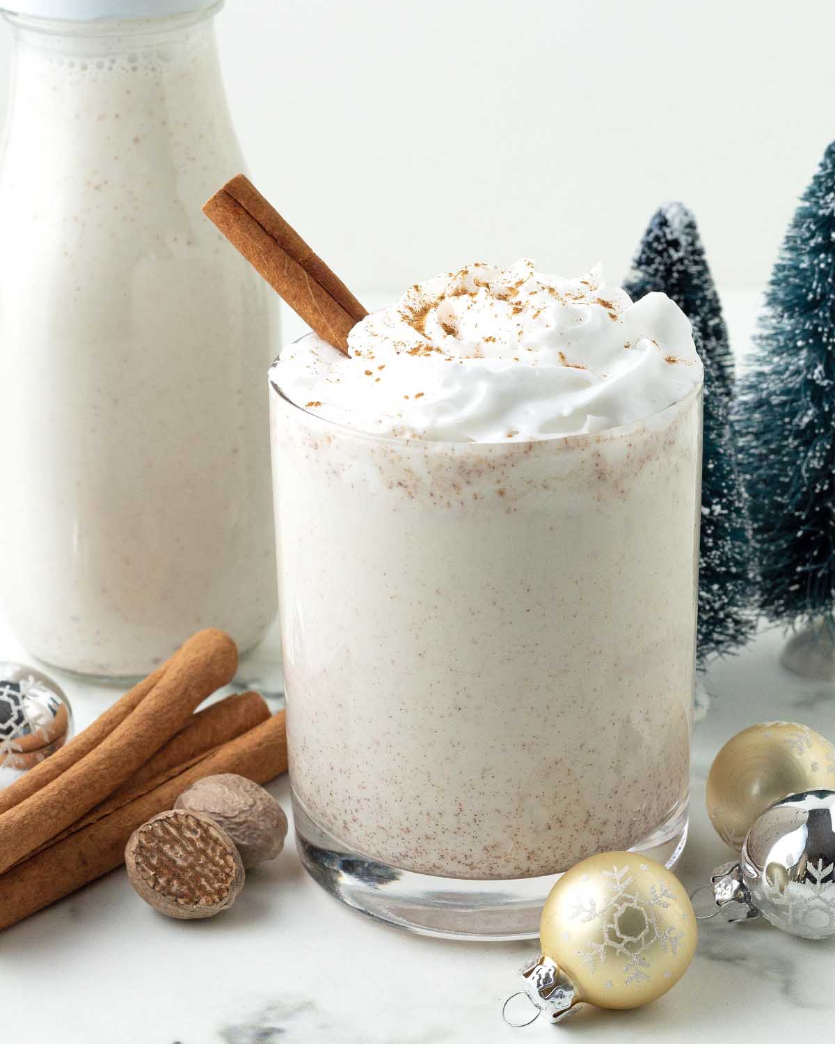A glass of vegan eggnog topped with coconut whipped cream, the glass is surrounded by holiday decorations and spices.