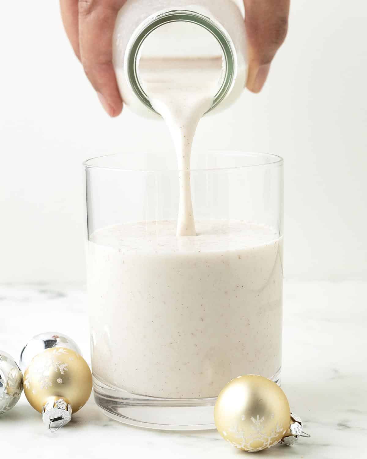 A hand pouring oat milk eggnog from a bottle into a glass