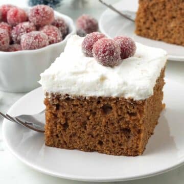 A square piece of gingerbread cake topped with frosting and sugared cranberries sitting on a small white plate with a fork on the side.