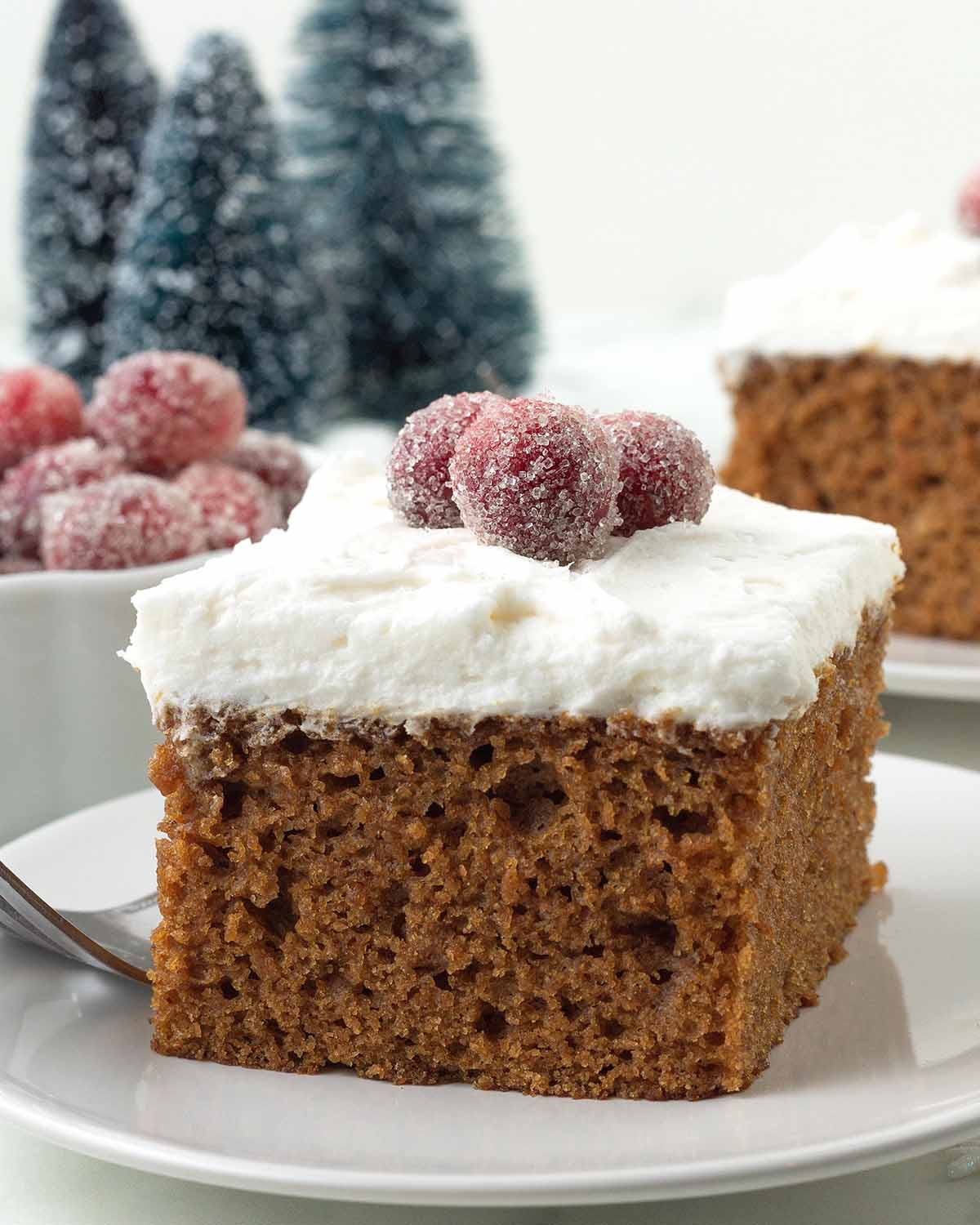 A square of vegan gingerbread cake on a plate, the cake is topped with frosting and sugared cranberries.