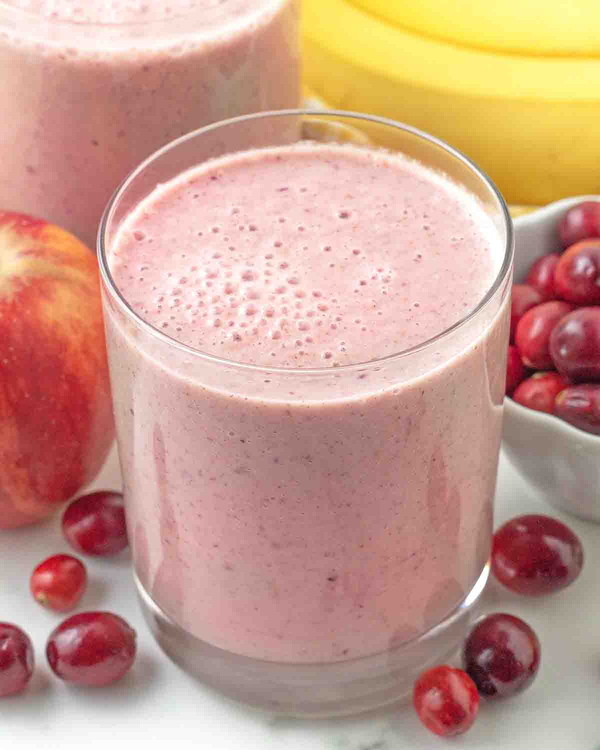 A glass of filled with cranberry smoothie, fresh cranberries, a fresh apple and bananas sit around the glass.