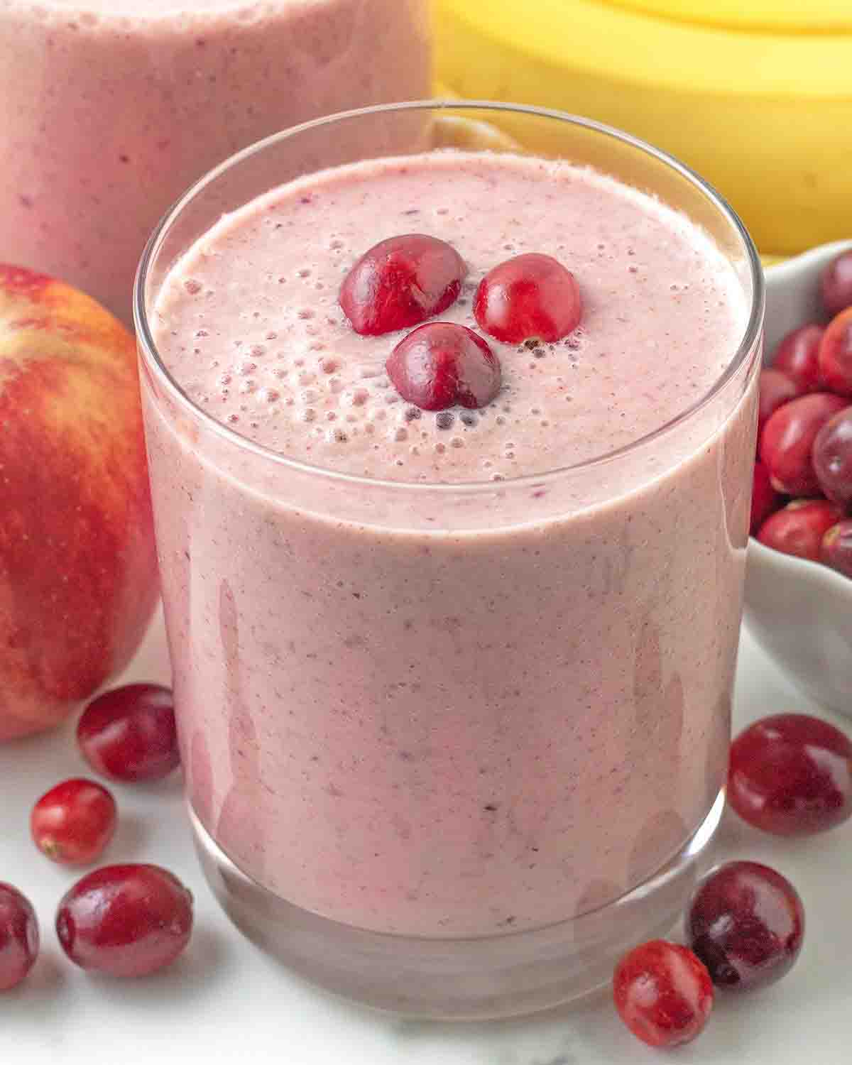 A glass of apple cranberry smoothie, the glass is surrounded by fresh cranberries.