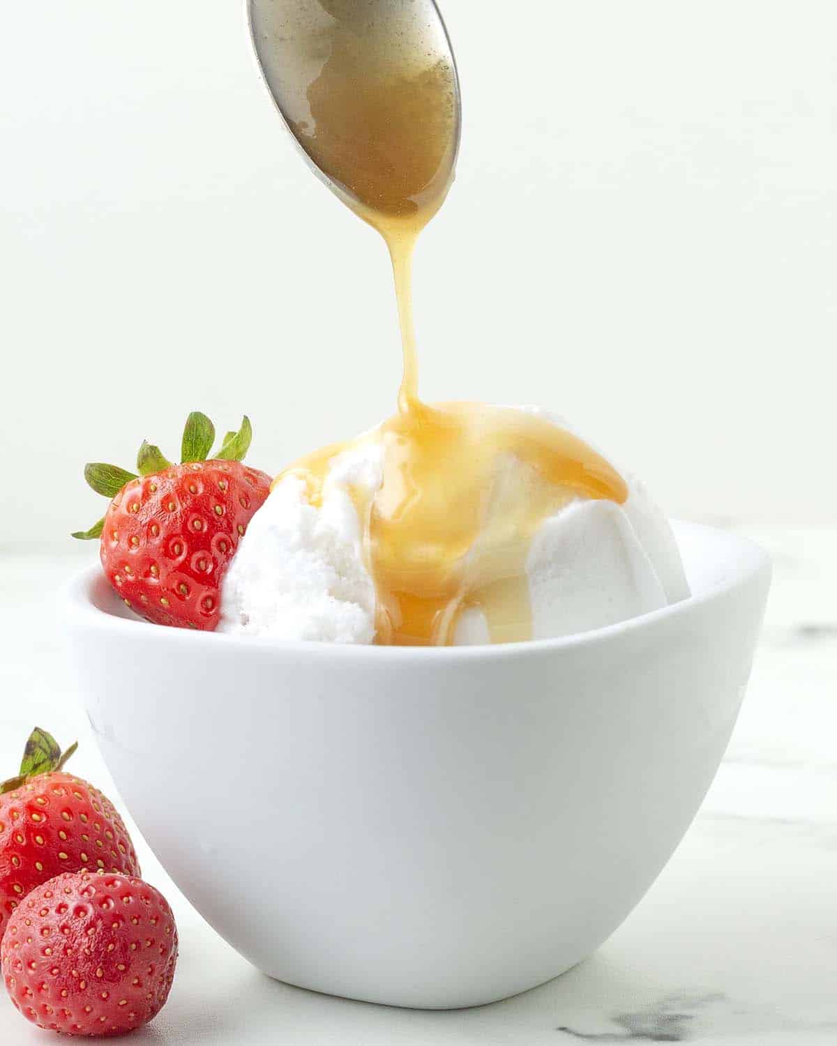 A spoon drizzling dairy-free maple caramel sauce onto coconut ice cream.