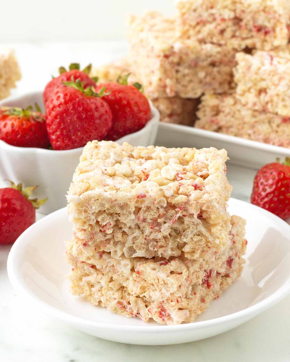 Two strawberry rice crispy treats on small plate, more treats and fresh strawberries sit in the background.