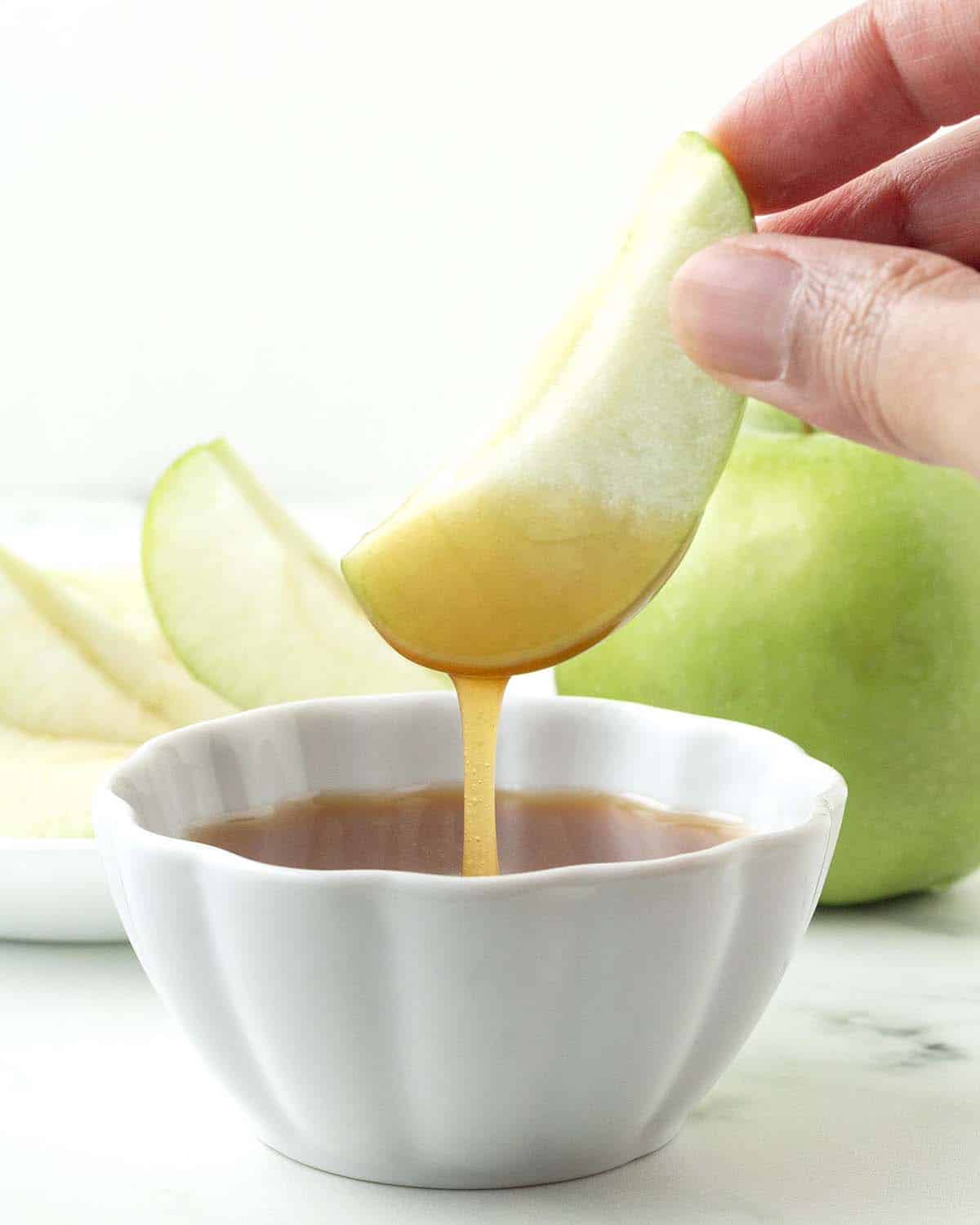 A hand dipping a slice of apple into a small bowl of maple caramel sauce.