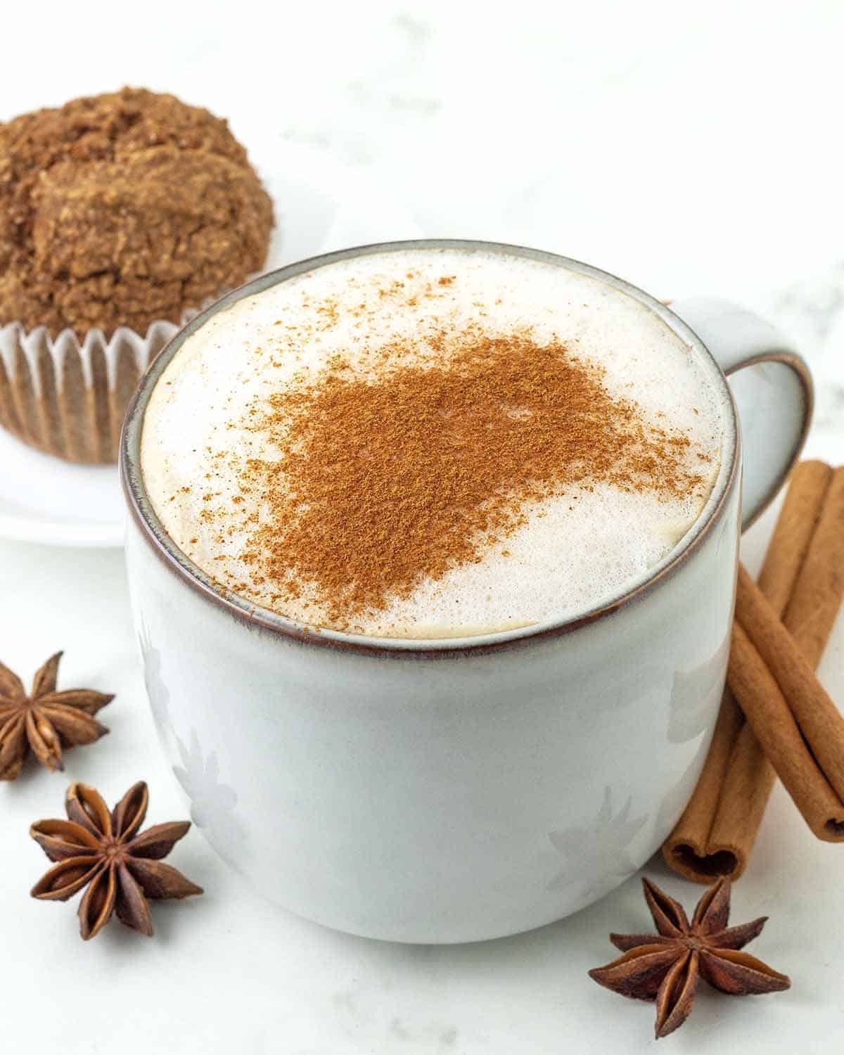 A vegan chai latte in a mug, a muffin sits behind the mug and dried spices sit around the mug.