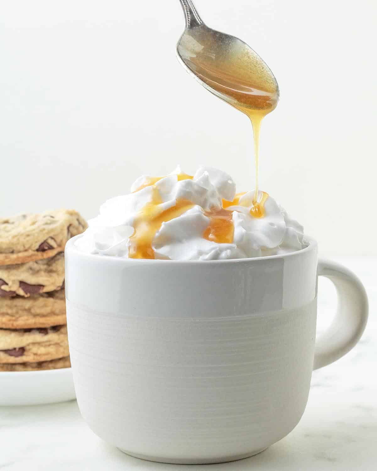 A spoon drizzling maple caramel onto a drink topped with whipped cream.