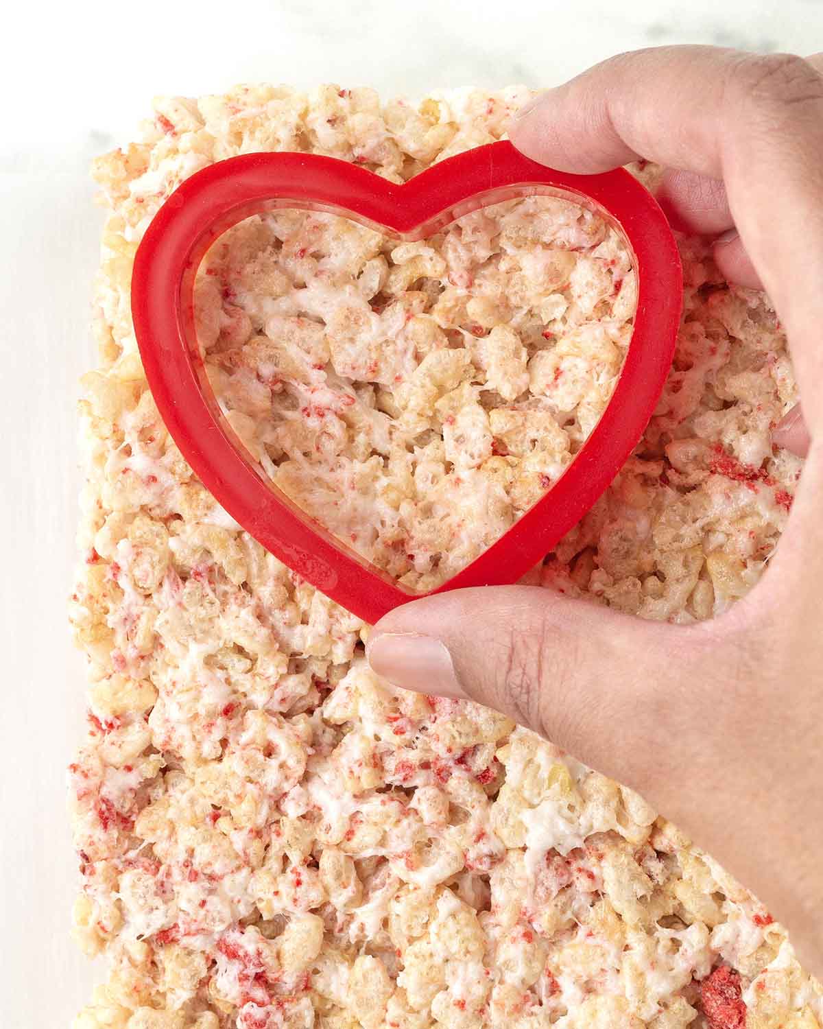 A hand pressing a heart shaped cutter into a square of rice crispy treats.