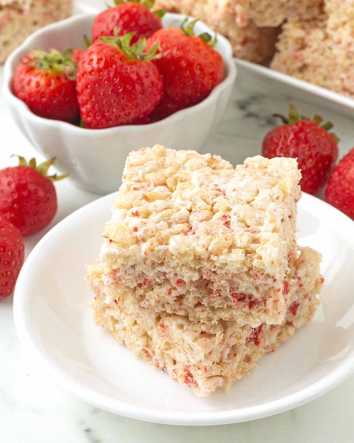 Two rice crispy treats on small plate, more treats and fresh strawberries sit in the background.