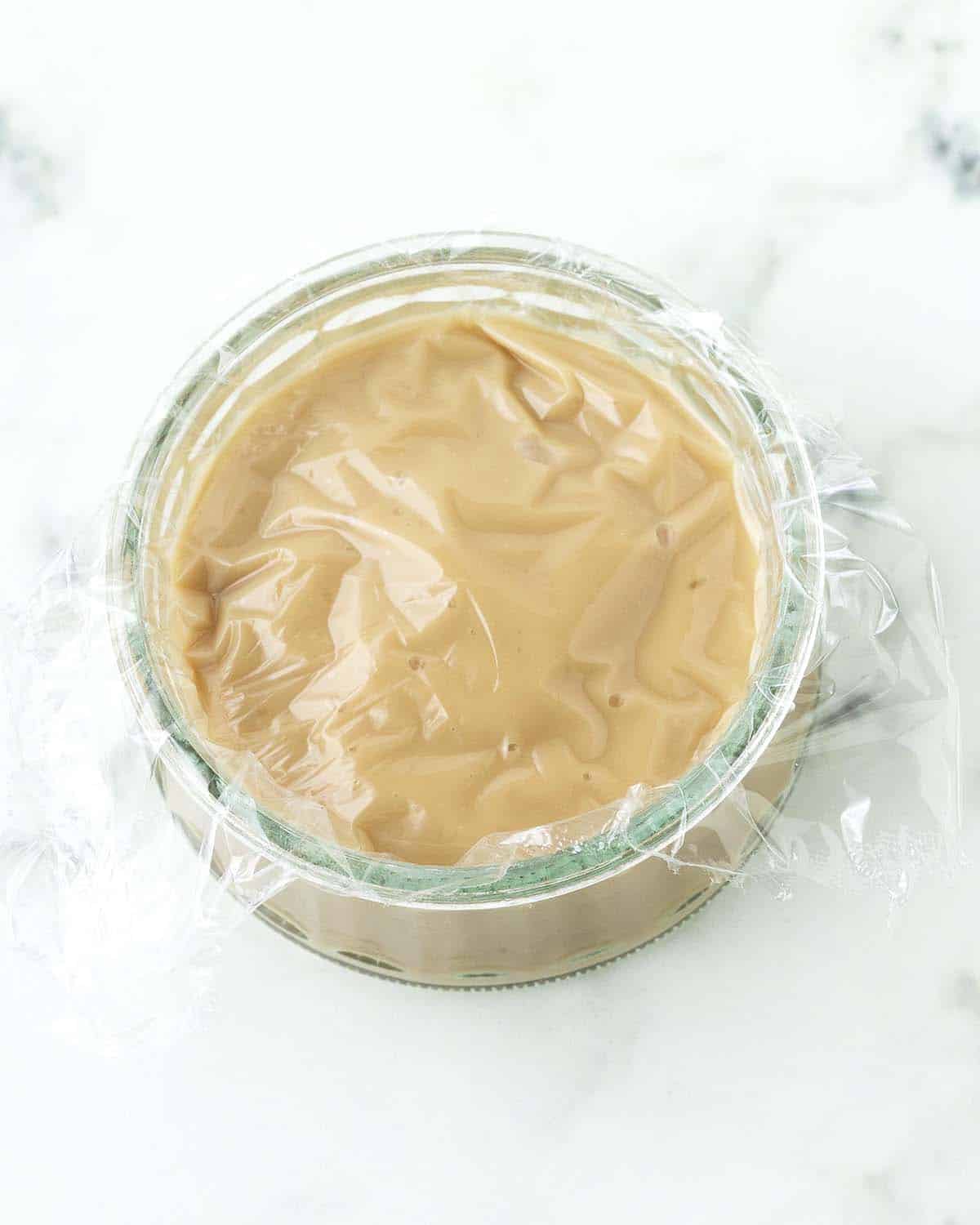 An overhead image of a small glass bowl filled with butterscotch pudding and covered in plastic wrap.