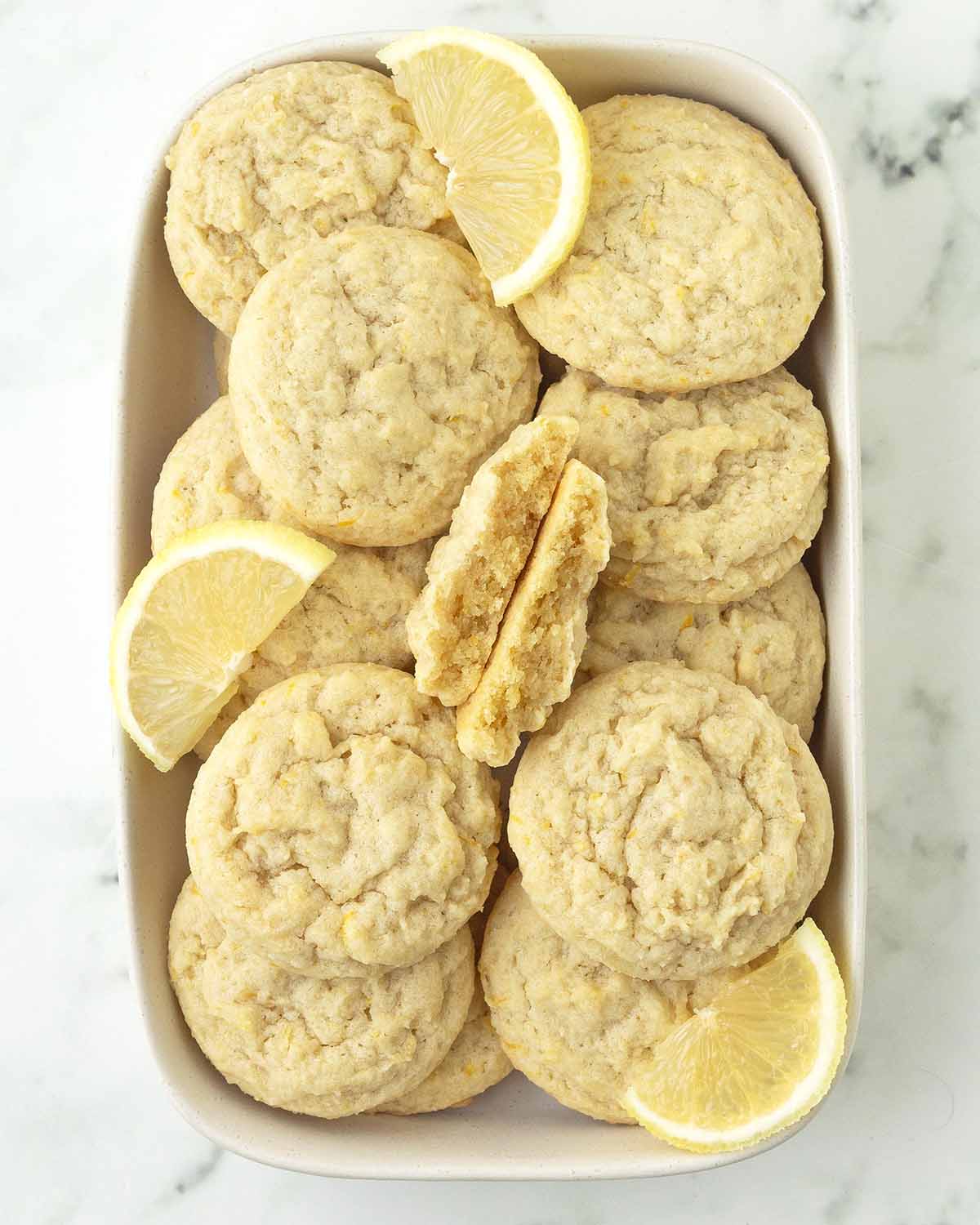 An overhead image of eggless lemon cookies in a dish, one cookie is split in half to show the soft inside.