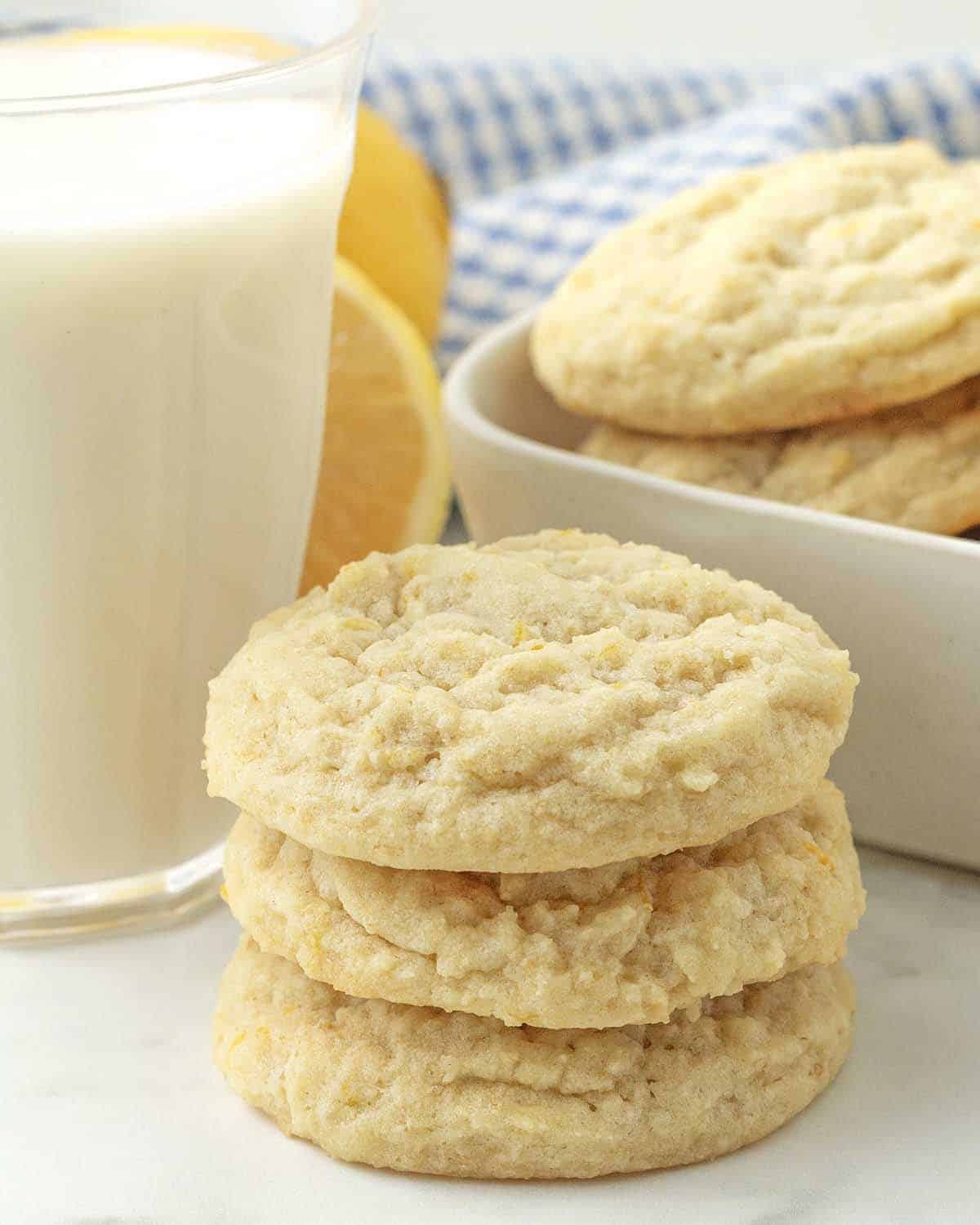 Three lemon cookies stacked on each other, a glass of milk and more cookies sit in the background.