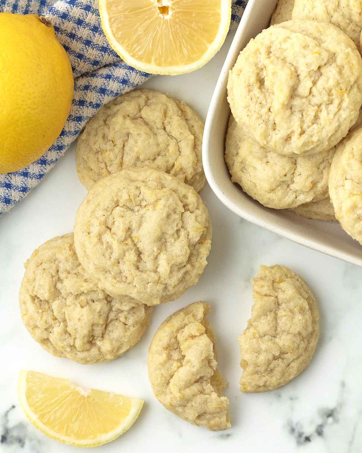 An overhead image showing lemon cookies and fresh lemons on a table, one cookie is broken in half.
