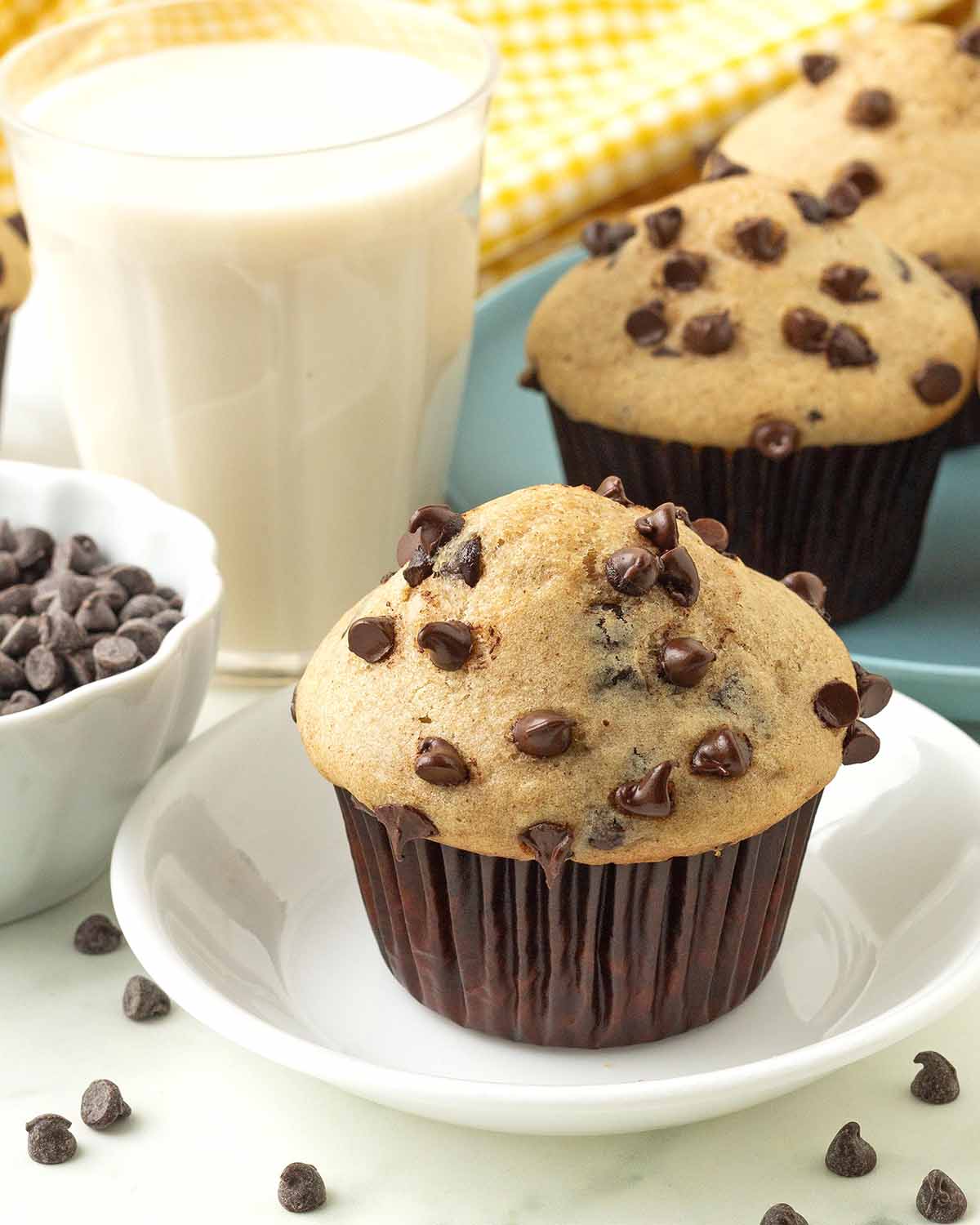 A chocolate chip muffin sitting on a white plate, more muffins and a glass of milk sit in the background.