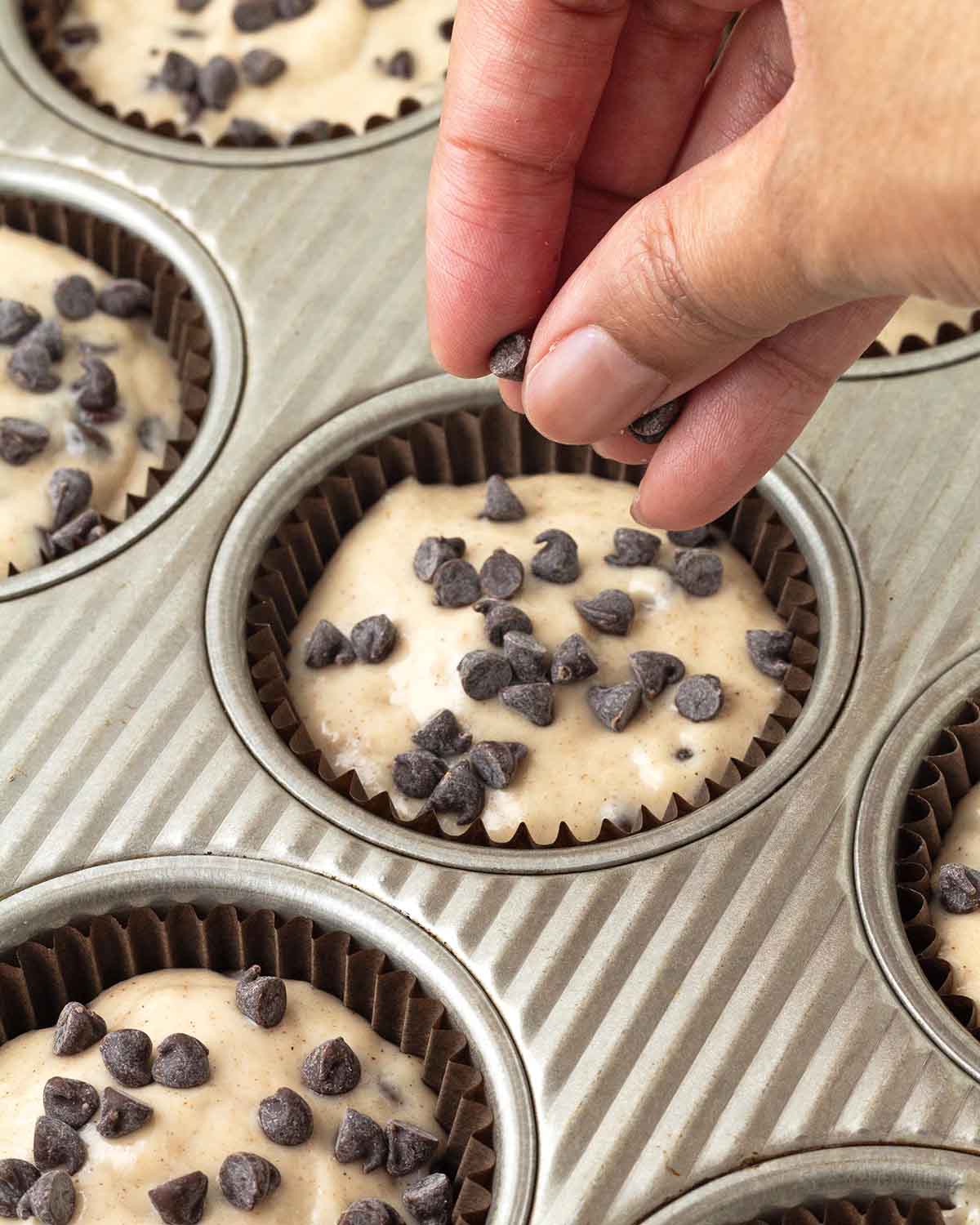 A hand sprinkling mini chocolate chips on top of an unbaked muffin.