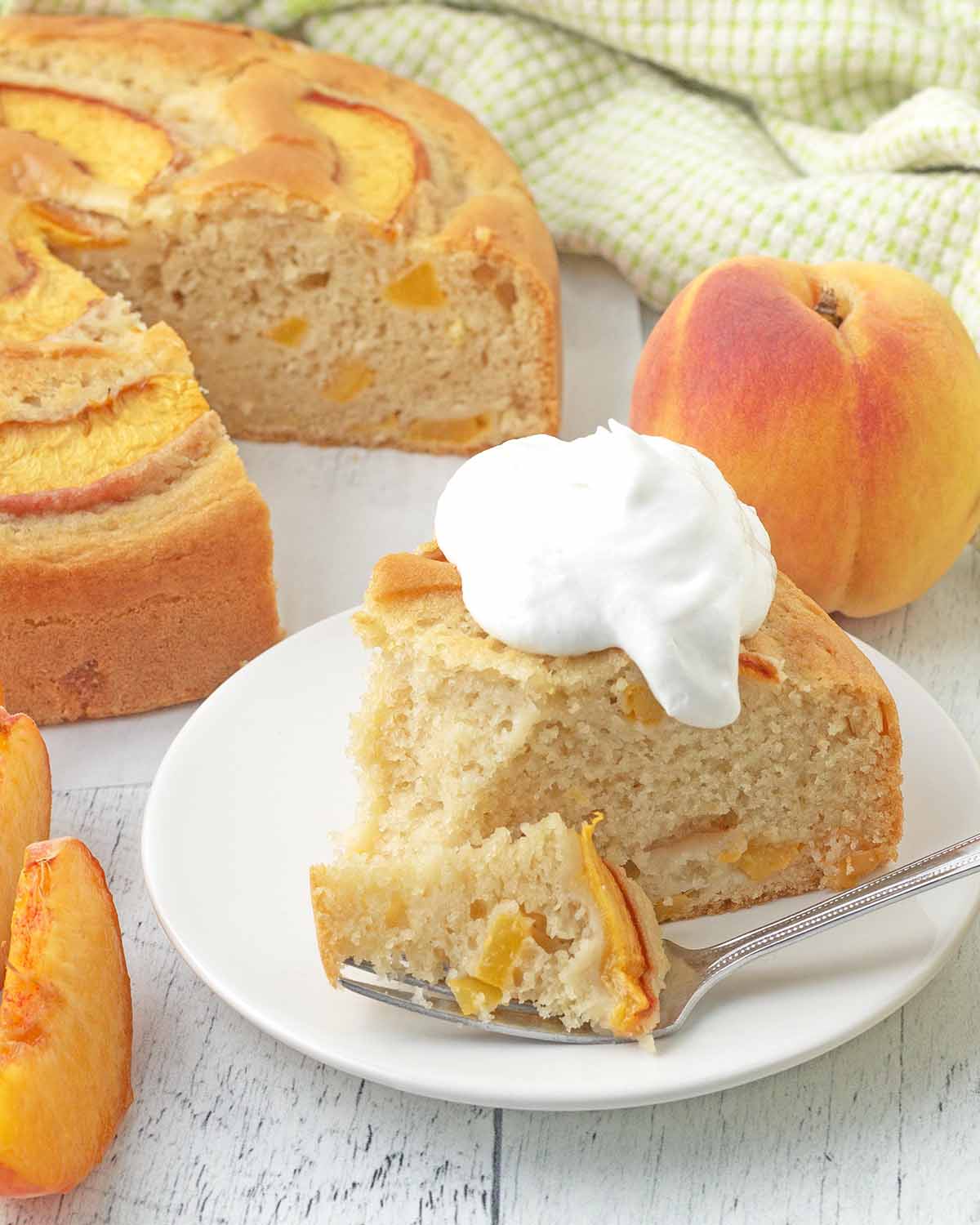 A slice of whipped cream topped peach cake on a white plate, a fork with a piece of the cake on it sits in the plate.