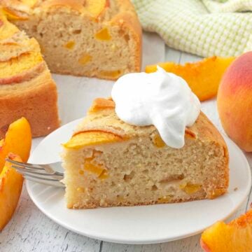 A slice of whipped cream topped peach cake on a white plate, a fork sits on the side of the cake.
