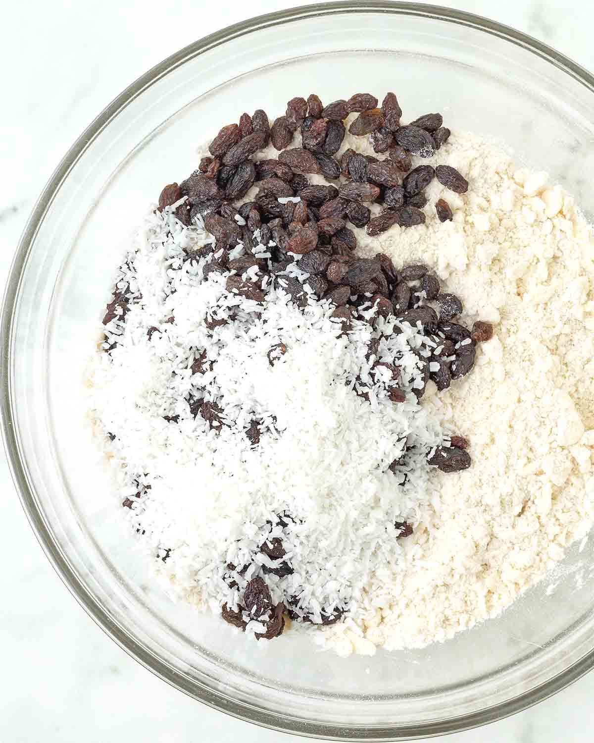 The dry ingredients for coconut scones in a glass bowl, the add-ins (shredded coconut and raisins) are on top.