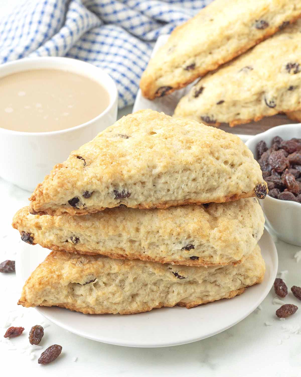 Three coconut scones stacked on a plate, more scones and a cup of tea sits behind the plate.