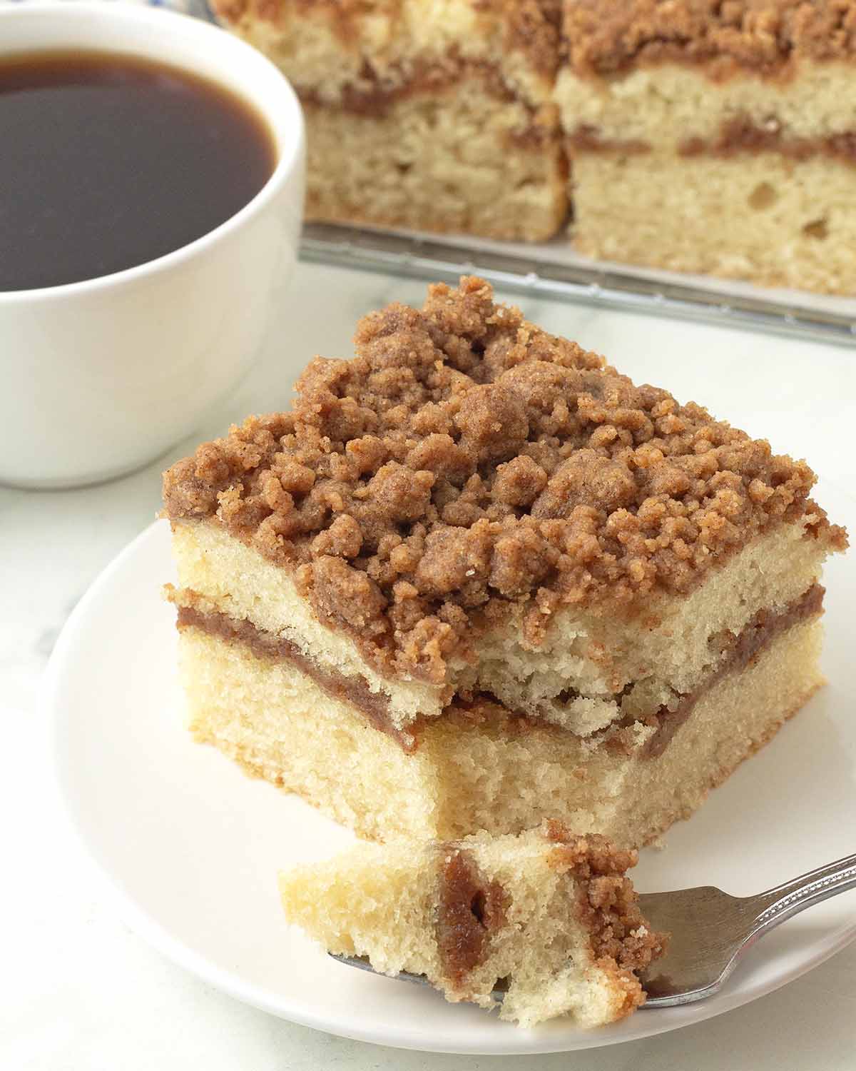 A piece of coffee cake on a white plate, a fork with a piece of the cake on it sits in front of the cake.