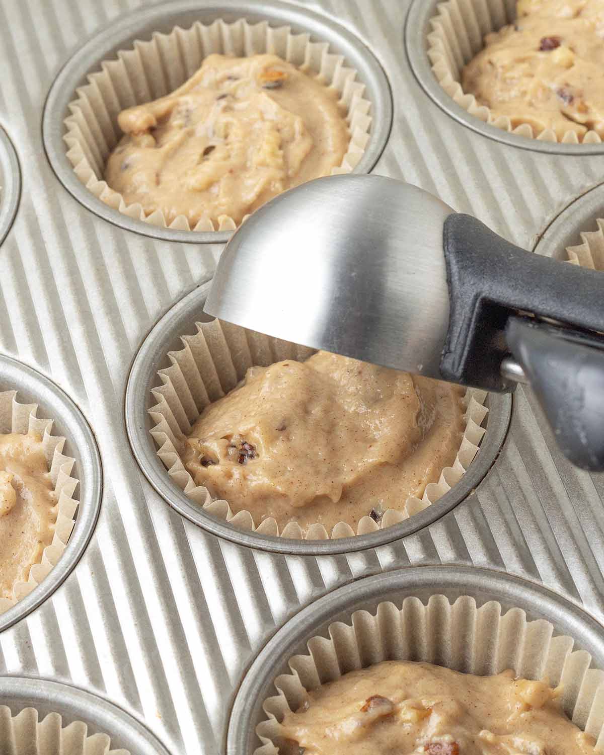 A medium cookie scoop adding banana muffin batter to a paper muffin cup in a muffin pan.