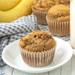 A banana muffin sitting on a white plate, more muffins, a glass of milk, bananas, and a dish cloth sit in the background.