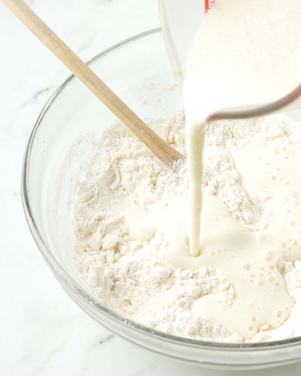 Vegan buttermilk being poured from a measuring cup into a glass bowl that holds the dry ingredients for biscuits.