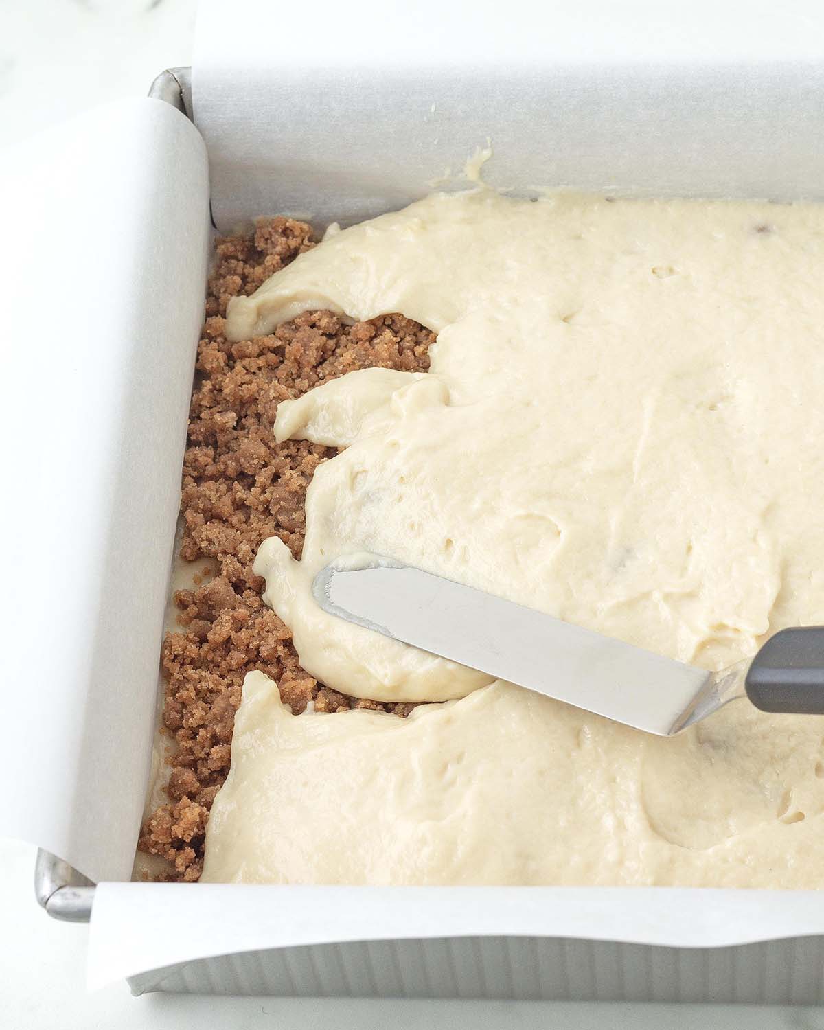 Batter being spread on top of a layer of cinnamon crumb in a baking pan.