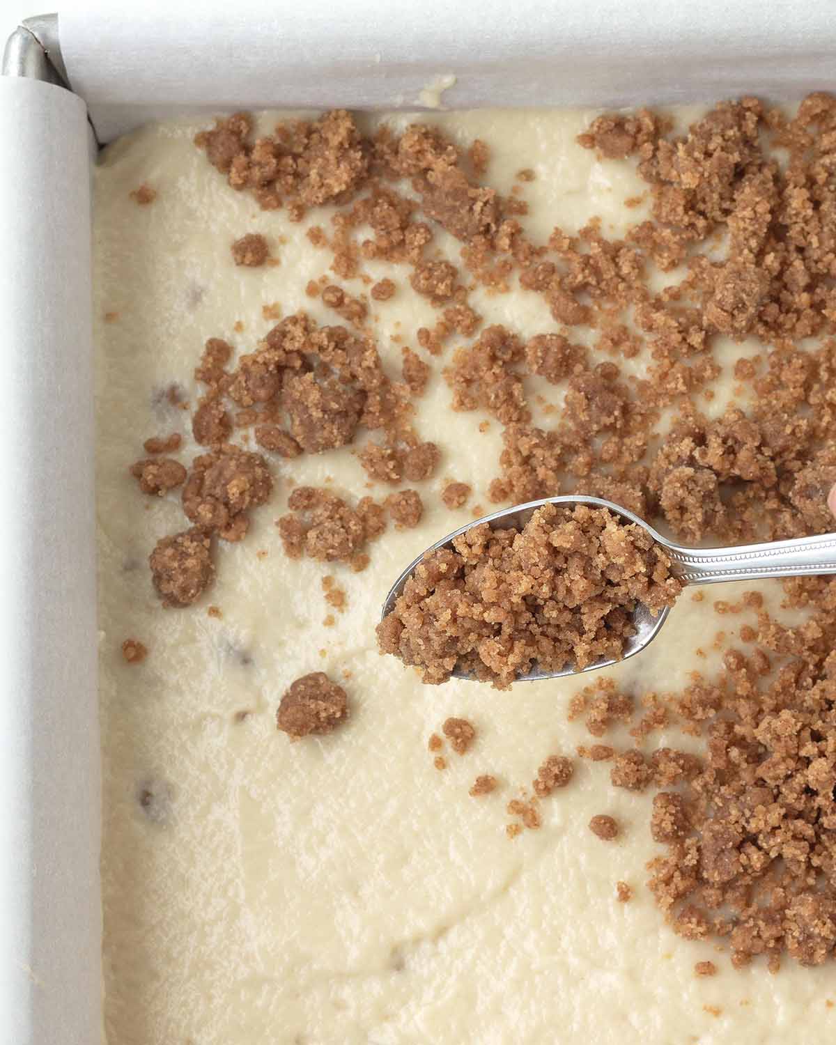 Cinnamon crumb being sprinkled from a spoon onto coffee cake batter that's in a baking pan.